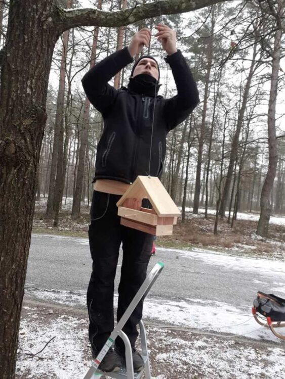 Andrej Smirnow zawiesza własnoręcznie wykonany karmnik dla ptaków na drzewie w parku w Mariampolu