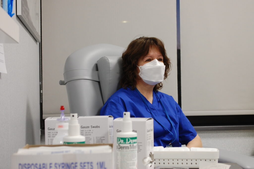 Lekarka w niebieskim fartuchu i masce ochronnej siedzi w gabinecie lekarskim