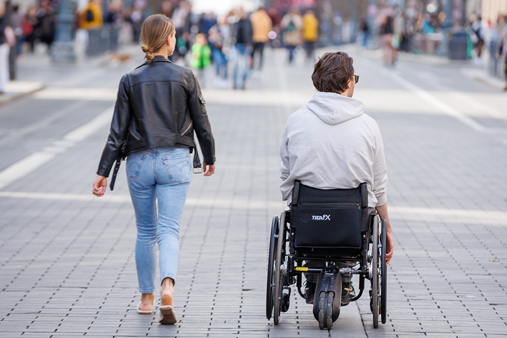 Kobieta idąca ulicą z mężczyzną na wózku inwalidzkim.
